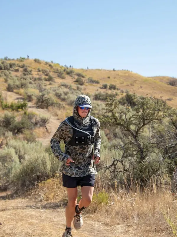Solo runner in camo gear pushing up a sagebrush-covered hill under blue sky